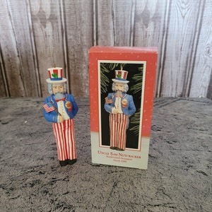 May include: A red, white, and blue Uncle Sam nutcracker ornament. The ornament is wearing a blue jacket, white pants with red stripes, a white hat with a red band, and a white beard. The ornament is holding a small American flag in one hand and a stick in the other. The ornament is dated 1988. The ornament is in its original box.