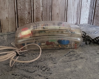 Könnte beinhalten: Ein transparentes, durchsichtiges Drehwahltelefon mit einem schwarzen Spiralkabel. Das Telefon hat einen roten Knopf und sichtbare interne Komponenten.