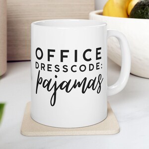 May include: White ceramic mug with the text "OFFICE DRESSCODE: pajamas" in black. The mug is on a beige coaster. The mug has a white handle and is in front of a wooden cutting board and a bowl of fruit.
