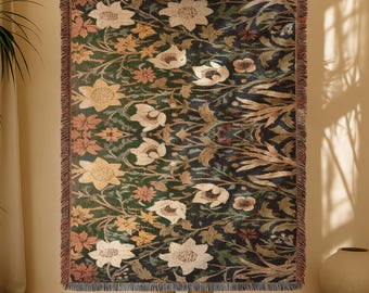 May include: A large, rectangular throw blanket with a floral pattern in shades of cream, gold, and rust against a dark green background. The blanket has a fringed edge and is held up by a person. The floral design includes large stylized flowers and foliage.