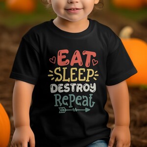 以下が含まれることがあります： マルチカラーのレトロなフォントで「EAT SLEEP DESTROY REPEAT」というテキストがプリントされた黒の半袖Tシャツ。デザインには小さなハートのアイコンと水平な矢印のグラフィックが含まれています。文字は4行にわたって配置されています。
