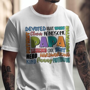 以下が含まれることがあります： 「PAPA」という言葉が大きくカラフルな文字で書かれた白いTシャツ。その周りには、「Devoted」、「Brave」、「Wise」、「Hero」、「Funny」などの言葉があります。このシャツはカジュアルなスタイルです。