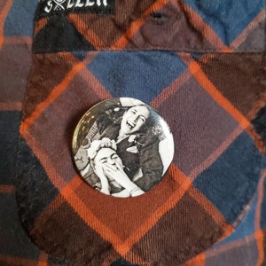 May include: A circular button with a black and white image of two people laughing, pinned to a plaid shirt. The shirt is a mix of orange, navy blue, and brown. The shirt has a pocket and a "Sullen" label.