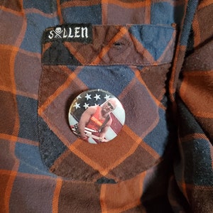 May include: A button pin featuring a patriotic design with an image of a wrestler, attached to a plaid flannel shirt. The shirt is primarily orange, black, and blue, with a pocket bearing the brand name "Sullen".