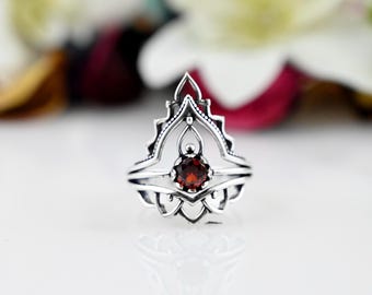 May include: A silver ring with a delicate, intricate design featuring a red gemstone in the center. The ring has a pointed, crown-like shape.
