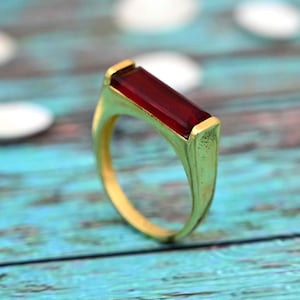May include: A gold-colored ring with a rectangular, deep red gemstone. The ring has a simple, elegant design with a smooth, polished finish. The gemstone is set in a bezel setting, and the band is slightly tapered.