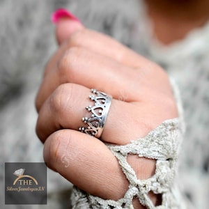 Può includere: Un anello d'argento con un design a corona. L'anello è indossato su un dito.