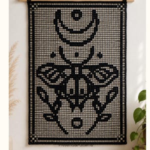 May include: A black macrame wall hanging featuring a grid pattern with a central moth design, a crescent moon, and circular motifs. The piece is suspended from a light-coloured wooden dowel with a black cord, creating a rectangular textile art display.