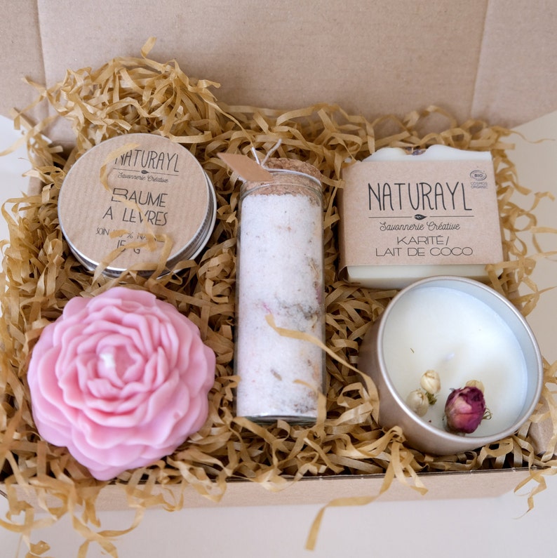 K&ouml;nnte beinhalten: Eine Geschenkbox gef&uuml;llt mit nat&uuml;rlichen Bade- und K&ouml;rperpflegeprodukten. Die Box enth&auml;lt eine rosa rosenf&ouml;rmige Kerze, ein kleines Glas Lippenbalsam, ein Glas mit Badesalz und eine Seife. Die Seife ist mit "NATURAYL Savonnerie Creative KARITE/ LAIT DE COCO" beschriftet.
