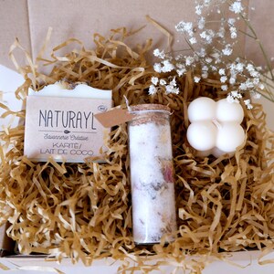 K&ouml;nnte beinhalten: Eine Geschenkbox gef&uuml;llt mit braunem Seidenpapier, die eine Seife mit der Aufschrift "NATURAYL Savonnerie Cr&eacute;ative KARIT&Eacute;/LAIT DE COCO", ein Glas mit Korkverschluss gef&uuml;llt mit Badesalz und getrockneten Blumen sowie eine wei&szlig;e Kerze mit vierbl&auml;ttrigem Design enth&auml;lt.