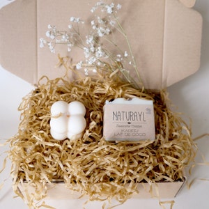 K&ouml;nnte beinhalten: Eine wei&szlig;e Geschenkbox gef&uuml;llt mit braunem Seidenpapier. Die Box enth&auml;lt eine wei&szlig;e Seife mit dem Text "NATURAYL Savonnerie Cr&eacute;ative KARIT&Eacute;/ LAIT DE COCO" und eine wei&szlig;e, runde, vierbl&auml;ttrige Kerze.