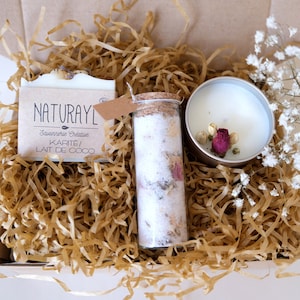 K&ouml;nnte beinhalten: Eine Geschenkbox gef&uuml;llt mit braunem Seidenpapier, die eine wei&szlig;e Seife mit dem Text "NATURAYL Savonnerie Cr&eacute;ative KARITE/ LAIT DE COCO", ein Glasgef&auml;&szlig; mit einem Korkverschluss gef&uuml;llt mit wei&szlig;en Badesalzen und getrockneten Blumen und eine wei&szlig;e Kerze mit getrockneten Blumen in einem silbernen Beh&auml;lter enth&auml;lt.