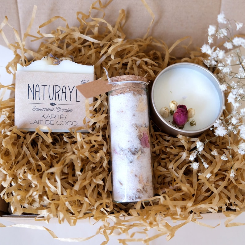 K&ouml;nnte beinhalten: Eine Geschenkbox gef&uuml;llt mit braunem Seidenpapier, die eine wei&szlig;e Seife mit dem Text "NATURAYL Savonnerie Cr&eacute;ative KARITE/ LAIT DE COCO", ein Glasgef&auml;&szlig; mit einem Korkverschluss gef&uuml;llt mit wei&szlig;en Badesalzen und getrockneten Blumen und eine wei&szlig;e Kerze mit getrockneten Blumen in einem silbernen Beh&auml;lter enth&auml;lt.