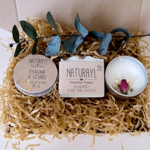 K&ouml;nnte beinhalten: Eine Geschenkbox gef&uuml;llt mit braunem Seidenpapier und drei Artikeln: ein kleines wei&szlig;es Glas mit braunem Deckel, beschriftet mit "NATURAYL Savonnerie Cr&eacute;ative BAUME A LEVRES 100% NATUREL 15 G", eine wei&szlig;e Seife, beschriftet mit "NATURAYL Savonnerie Cr&eacute;ative KARITE/ LAIT DE COCO", und eine kleine wei&szlig;e Kerze mit silbernem Deckel und getrockneten rosa Bl&uuml;ten im Inneren.