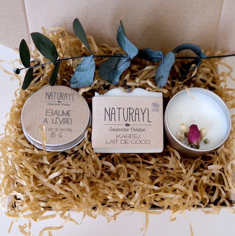 K&ouml;nnte beinhalten: Eine Geschenkbox gef&uuml;llt mit braunem Seidenpapier und drei Artikeln: ein kleines wei&szlig;es Glas mit braunem Deckel, beschriftet mit "NATURAYL Savonnerie Cr&eacute;ative BAUME A LEVRES 100% NATUREL 15 G", eine wei&szlig;e Seife, beschriftet mit "NATURAYL Savonnerie Cr&eacute;ative KARITE/ LAIT DE COCO", und eine kleine wei&szlig;e Kerze mit silbernem Deckel und getrockneten rosa Bl&uuml;ten im Inneren.