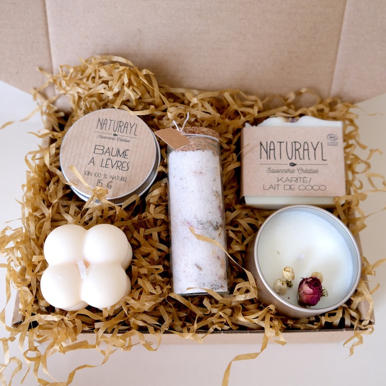 K&ouml;nnte beinhalten: Eine Geschenkbox gef&uuml;llt mit nat&uuml;rlichen Sch&ouml;nheitsprodukten. Die Box enth&auml;lt ein kleines Glas Lippenbalsam mit der Aufschrift "NATURAYL Baume a Levres 15g", eine Glasflasche mit K&ouml;rperscrub, eine Seife mit der Aufschrift "NATURAYL Savonnerie Creative Karite/ Lait de Coco" und eine kleine Kerze mit getrockneten Blumen.
