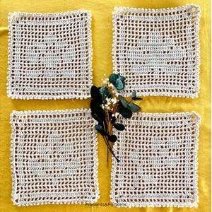 Può includere: Quattro centrini quadrati all'uncinetto color bianco sporco con un motivo traforato a griglia e un bordo smerlato. I quattro pezzi sono disposti a quadrante con un piccolo rametto di foglie di eucalipto verde scuro al centro.