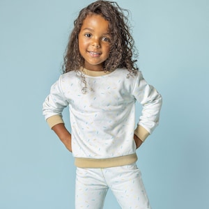 May include: A young girl wearing a white sweatshirt and matching pants with a sprinkle pattern. The sweatshirt has a tan ribbed collar and cuffs. The pants have a tan waistband. She is wearing pink and blue tie-dye sneakers.