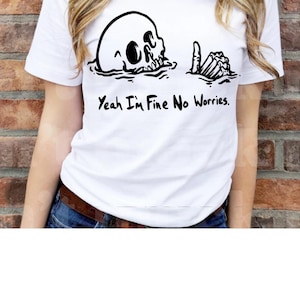 Pode incluir: Camiseta branca com um gráfico preto de uma caveira na água e uma mão de esqueleto levantando o polegar. O texto "Yeah I'm Fine No Worries" está impresso abaixo. A camiseta é usada por uma pessoa.