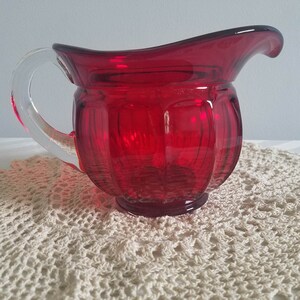 May include: A red glass creamer with a clear glass handle. The creamer has a ribbed design and is sitting on a white crocheted doily.