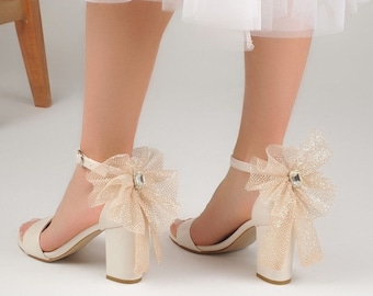 Può includere: Sandali bianchi con tacco in raso con un grande fiocco beige scintillante sul retro di ogni scarpa.