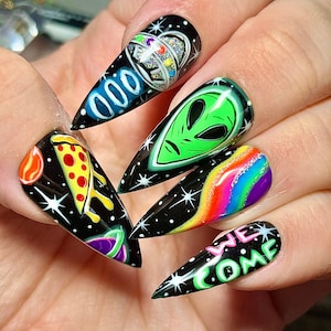 May include: Close-up of a hand with long, black stiletto nails. Each nail is painted with a different design, including a UFO, an alien face, a pizza slice, a rainbow, and the words "We Come".