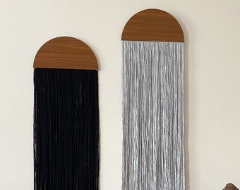 May include: Two wall hangings with wooden half-circle tops and long strands of yarn hanging down. The yarn is black on the left and gray on the right.
