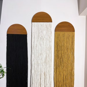 May include: Three wall hangings with wooden half-circle tops and long strands of yarn hanging down. The yarn is black, white, and mustard yellow.