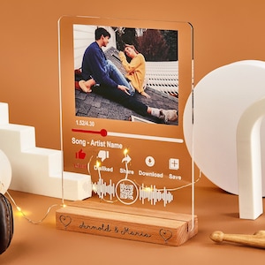 May include: A clear acrylic photo frame with a couple's photo and a music player interface. The frame has a wooden base with the names "Arnold & Maria" engraved on it.
