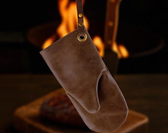May include: A brown leather oven mitt with a loop for hanging. The mitt has a classic design with a thumb and a separate space for the fingers. The background shows a blurred image of a grill and a wooden cutting board.