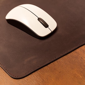 May include: A white wireless computer mouse with a black scroll wheel rests on a brown leather mouse mat. The mouse mat has stitched edges and sits on a wooden surface.