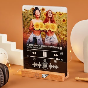 May include: A glass photo frame with a picture of two friends in a field of sunflowers. The frame has a black background with a music player interface and the text "A good friend is cheaper than therapy" and "Topeka, Kansas, 2021".