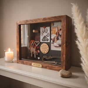 May include: A rectangular wooden shadow box memorial display case with a dark brown finish. The front features a brass plaque engraved with the name BUSTER. Inside are two photographs of a beagle, a paw print, a heart pendant, and a collar.