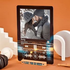 May include: A black and white photo of a couple in winter clothing is displayed on a wooden stand. The stand has a music player interface with a song title, artist name, and a QR code. The stand has a wooden base with the text "I LOVE YOU SO MUCH" printed on it.