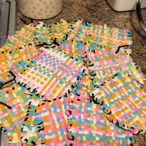 May include: A stack of colorful woven coasters with black trim. The coasters are made with a variety of bright colors, including pink, blue, yellow, and green.