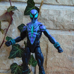 May include: Action figure of a superhero wearing a black and purple suit with green accents. The figure has a detailed sculpt and is posed in a dynamic stance.