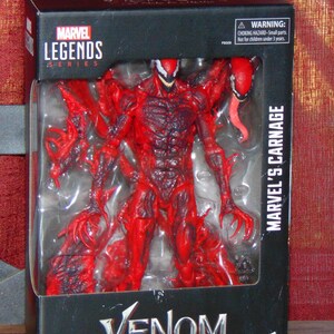 May include: A Marvel Legends Series action figure of Marvel's Carnage, packaged in a black box. The figure is predominantly red with black accents, featuring a detailed sculpt and a long, red tongue. The box reads "Venom: Let There Be Carnage."