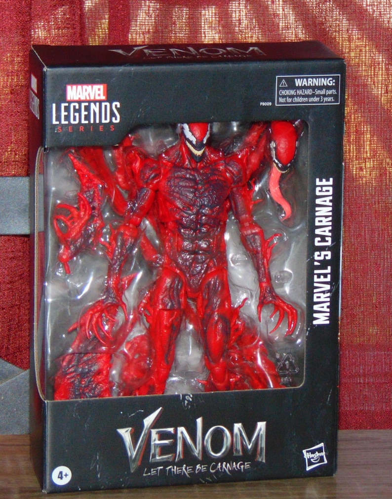 May include: A Marvel Legends Series action figure of Marvel's Carnage, packaged in a black box. The figure is predominantly red with black accents, featuring a detailed sculpt and a long, red tongue. The box reads "Venom: Let There Be Carnage."