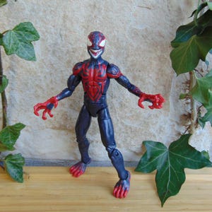 May include: A blue and red action figure of a character with a menacing grin. The figure has a red spiderweb design on the chest and arms, with red claws and feet. The figure is posed with arms outstretched.