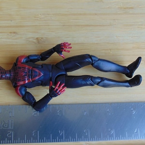 May include: A Miles Morales Spider-Man action figure in a dynamic pose, featuring a black and red suit. The figure is lying on a wooden surface next to a metal ruler. The figure's design includes web detailing.