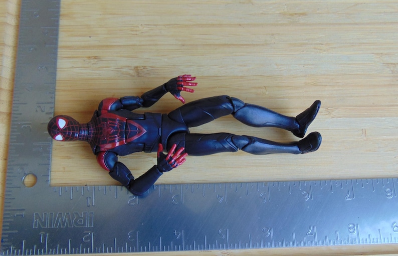 May include: A Miles Morales Spider-Man action figure in a dynamic pose, featuring a black and red suit. The figure is lying on a wooden surface next to a metal ruler. The figure's design includes web detailing.