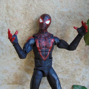 May include: A Miles Morales Spider-Man action figure in a dynamic pose. The figure features a black and red suit with a spider emblem on the chest. The figure's hands and mask details are red, with white eye lenses.