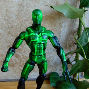 May include: A green action figure with a detailed design. The figure is primarily green with black accents on the limbs and a pattern of lighter green lines. The figure is posed with arms extended.