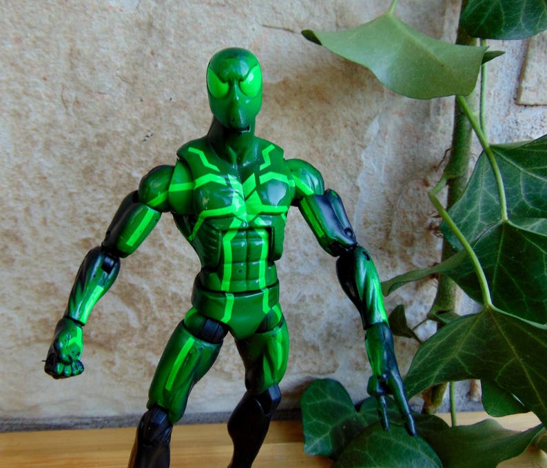 May include: A green action figure with a detailed design. The figure is primarily green with black accents on the limbs and a pattern of lighter green lines. The figure is posed with arms extended.