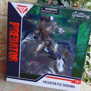 Op de afbeelding: Een Predator PVC diorama figuur in een display doos. De figuur staat in een gevechtshouding, met gedetailleerde bepantsering en wapens. De doos heeft het woord "Predator" in grote oranje letters en een waarschuwingslabel.