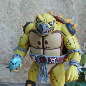 May include: A yellow action figure of a turtle-like character with a stern expression. The figure has a tan chest, a belt with skull and crossbones, and a blue weapon in its hand. The figure is approximately 6 inches tall.