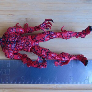 May include: A red and black action figure of a character with a white face and black accents. The figure is lying on a wooden surface next to a metal ruler. The figure's pose suggests movement.