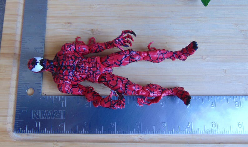 May include: A red and black action figure of a character with a white face and black accents. The figure is lying on a wooden surface next to a metal ruler. The figure's pose suggests movement.