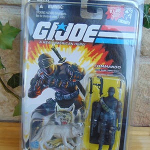 Pode incluir: Figura de ação G.I. Joe na embalagem original. A embalagem apresenta um gráfico do personagem Snake Eyes com um lobo e o texto "A Real American Hero". A figura é um Comando, nome de código Snake Eyes.