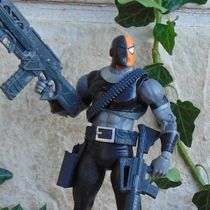 Peut inclure: Une figurine articulée Deathstroke avec un masque noir et orange, une combinaison tactique noire et grise, et une cartouchière. La figurine tient un fusil gris dans sa main droite et possède un second fusil fixé à sa hanche.
